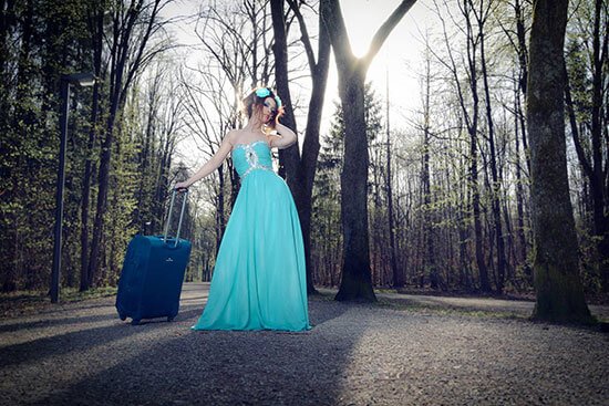 Eine junge Frau im Kleid und mit Reisetrolley steht mitten im Wald.