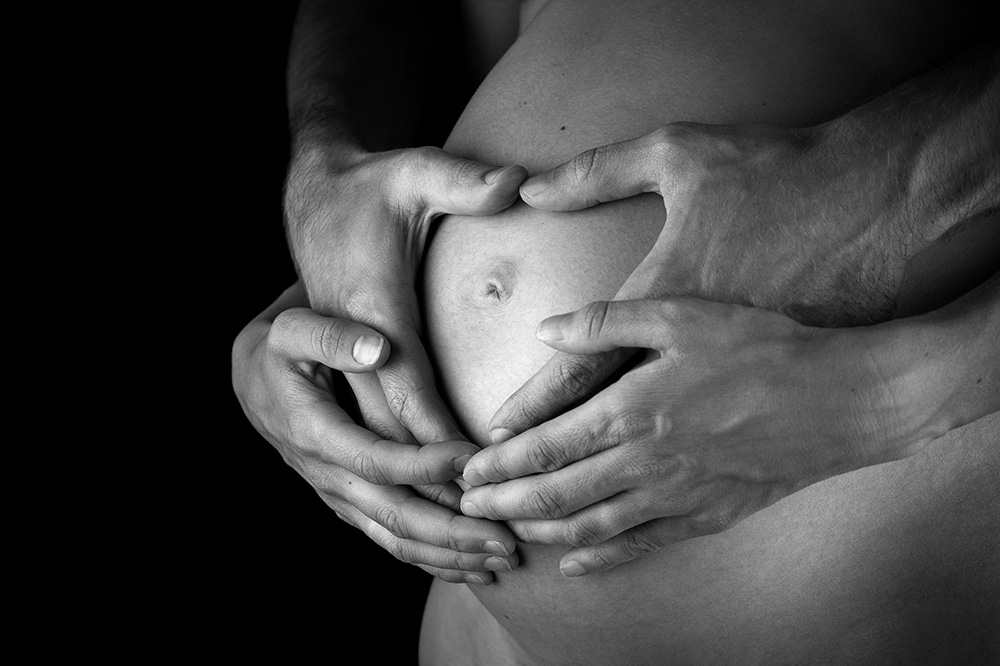 Vier Hände umarmen Babybauch und bilden eine Herzform